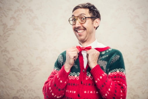 A man in a knit reindeer Christmas cardigan button up sweater, complete with matching red bow tie and a classy mustache.  He straightens his bow tie with a cheesy smile on his face, proud of his fashion style.  Damask style vintage wall paper in the background.  Horizontal with copy space | Image: Unsplash