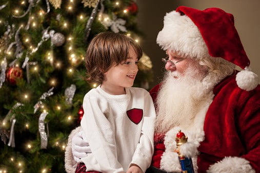 A young boy sits on Santa Claus' lap by a decorated Christmas tree in his living room. Santa has a real beard and the child looks at him with wonder, awe, and a hint of excitement | Image: Unsplash
