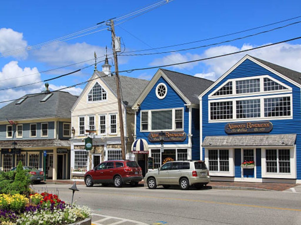 The image shows Dock Square lined with stores and restaraunts in Kennebunkport, Maine, New England. Street, sidewalk, flowers, American flag, parked cars and clear blue sky with some clouds.