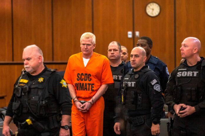 Alex Murdaugh standing in a court room with his prison wear in handcuffs and surrounded by police.