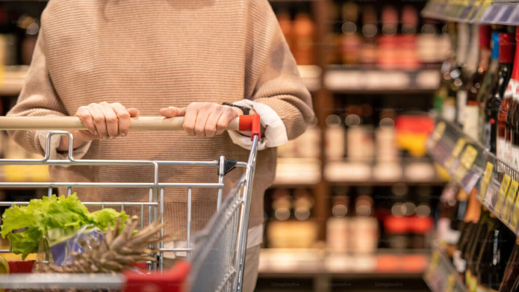 A picture of hands of female-consumer-in-beige-pullover-pushing-shopping-cart-with-fresh-food-products while walking along shelves with alcohol