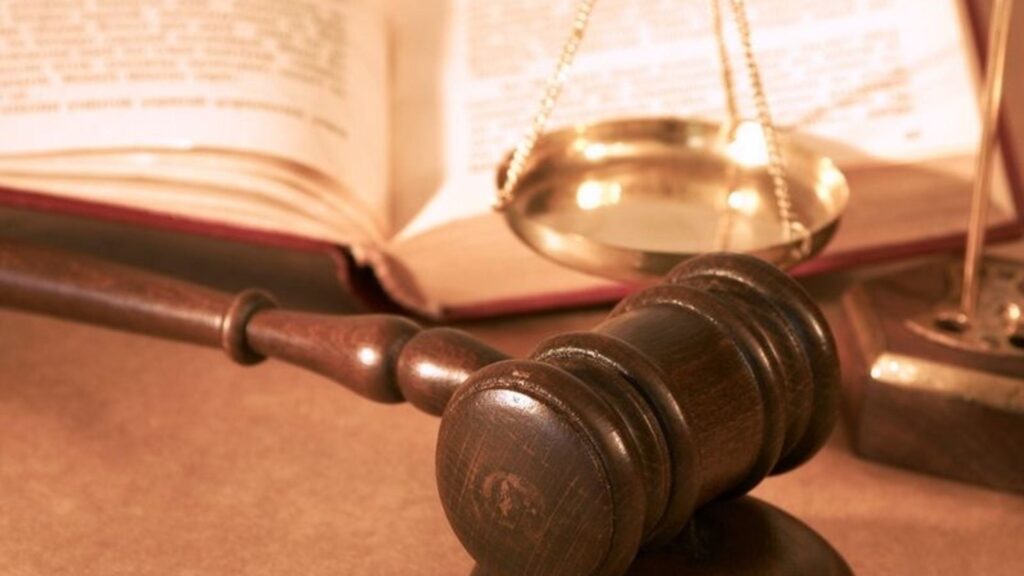 A gavel resting with a book and a scale in the backdrop.