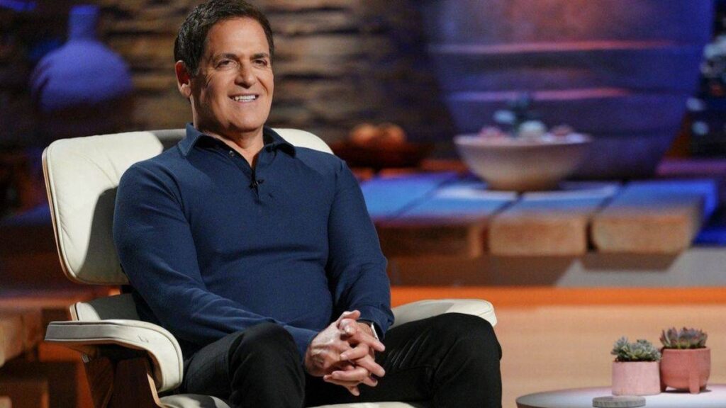 Mark Cuban is sitting in a chair with a smile.