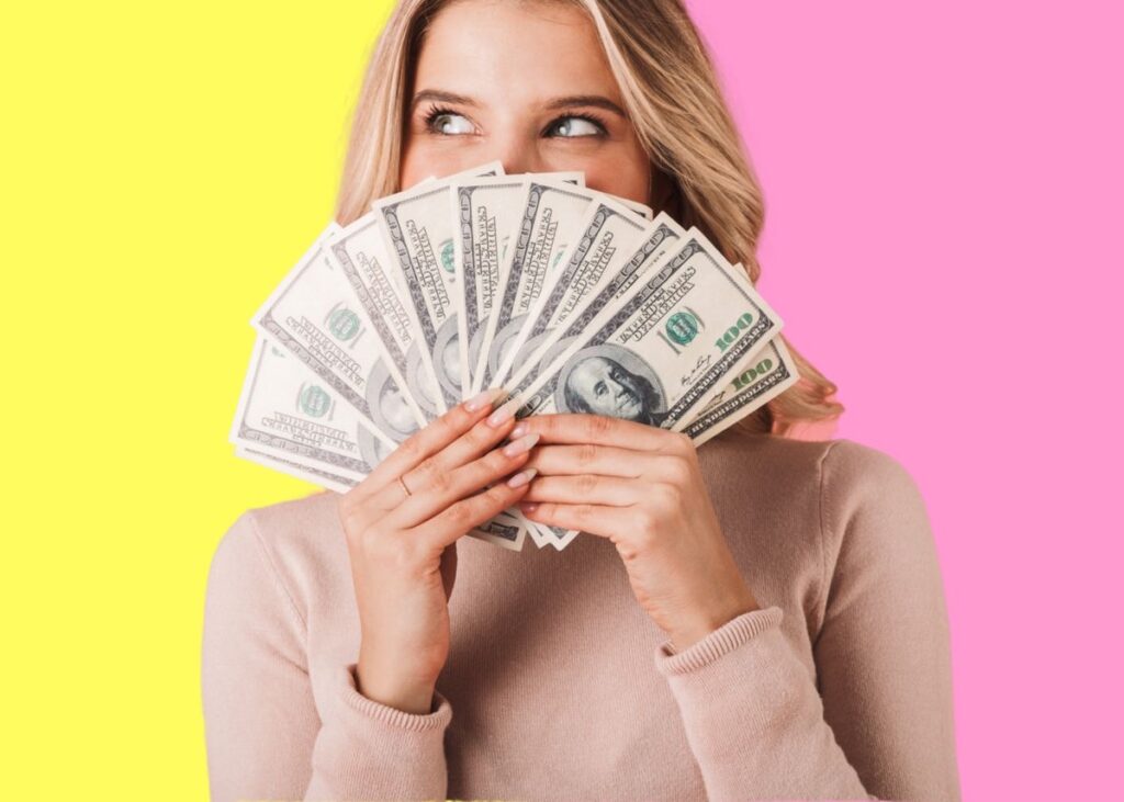 A lady holding several $100 bills spread across her face with a yellow and pink backdrop.
