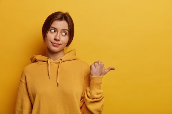 An unimpressed woman wearing a yellow hoodie and standing in front of a yellow background