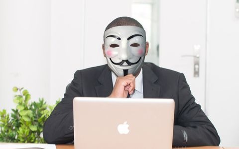 A man wearing an anonymous mask sitting behind a laptop