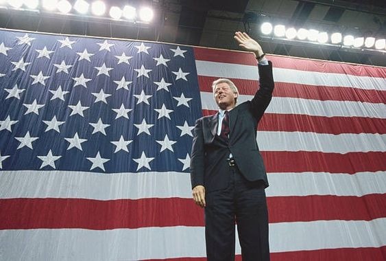 Bill Clinton standing in front of a large US flag waving to an audience