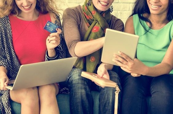 Three women of different races sitting next to eachother smiling and holding different devices