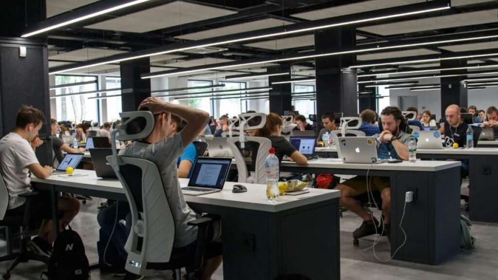 A wide-angle view of a bustling open-plan office filled with individuals working at their desks. Many are focused on laptops, with some appearing in mid-conversation or deep thought
